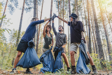 Multiracial group of volunteers taking care of nature ecology and picking garbage in plastic bags cleaning ground in forest and giving high five for teamwork celebrating it