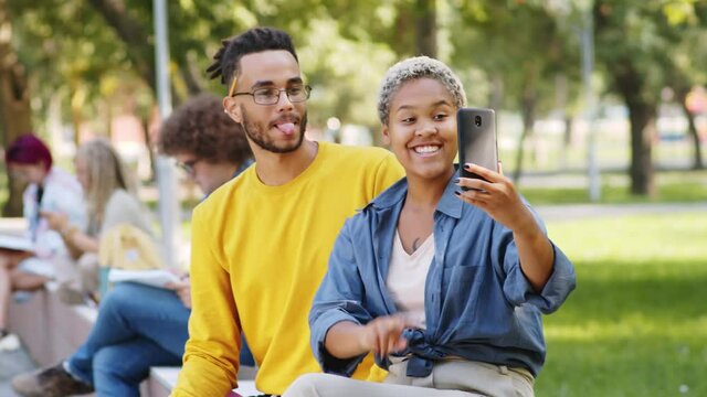 Positive mixed-race male and female college students smiling and posing together for smartphone camera while taking a selfie in park on summer day