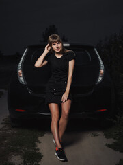 Fototapeta na wymiar Full body portrait of young blonde woman in black dress sneakers posing near electric zero emissions car twilight outdoors country road