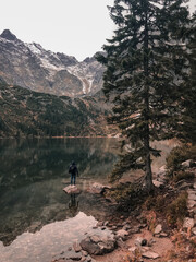 a young man a guy in a hood stands on the Big Stone in the middle of a clear azure green lake surrounded by high mountains and trees. Snow-covered tops of mountains and dry conifers in the fog.