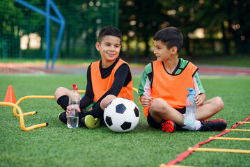 Two happy teen boys in football uniforms having a rest on the sport field and enjoying fresh water...