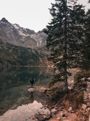 a young man a guy in a hood stands on the Big Stone in the middle of a clear azure green lake surrounded by high mountains and trees. Snow-covered tops of mountains and dry conifers in the fog.