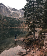 a young man a guy in a hood stands on the Big Stone in the middle of a clear azure green lake surrounded by high mountains and trees. Snow-covered tops of mountains and dry conifers in the fog.