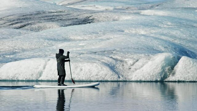 Pan right, person paddleboards through Iceland icebergs