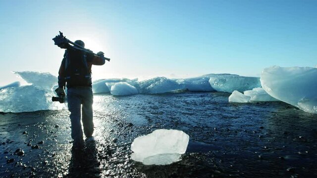Slow motion, man with tripod walks on Okulsarlon Beach covered in ice
