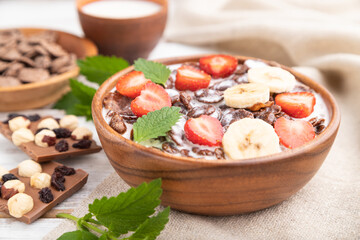 Chocolate cornflakes with milk and strawberry in wooden bowl on white wooden background. Side view, selective focus.