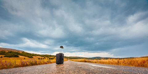 Suitcase with cap on empty road. Nature landscape and stormy sky. New journey, adventure and travel concept. Copy space