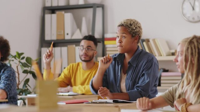Group of young multiethnic students sitting at desks in classroom and raising arms, then young mixed-race woman asking question and having discussion with professor during college class
