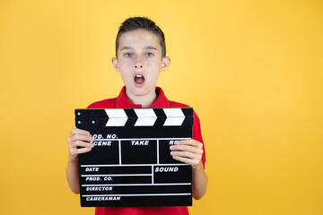 Young beautiful child boy over isolated yellow background surprised holding clapperboard