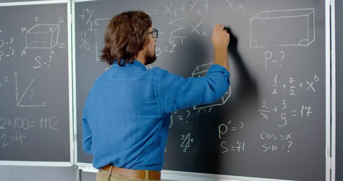 Rear of Caucasian male lecturer writing math or physics formulas with chalks on blackboard. Back view on man teacher working at school. Lesson of mathemathics. Portrait of mathematician.