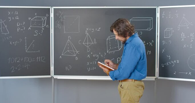 Caucasian male mathematician in glasses at backboard with math or physic formulas and laws writing notes in notebook. Man teacher in school checking equations of algebra or geometry with handbook.