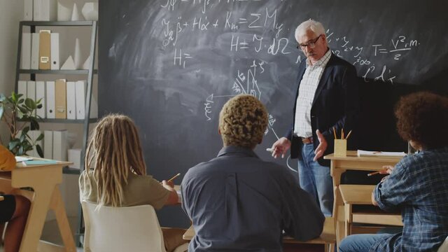 Senior professor pointing at formulas on chalkboard and explaining them to group of college students while giving lecture