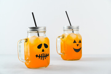 Iced pumpkin cocktails in glass jars decorated with scary faces on the gray table, white background. Halloween Party mocktails. Copy space