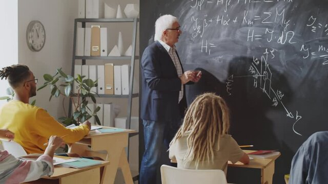 Elderly professor writing physics formulas on chalkboard and explaining them to group of young students while giving lecture in college