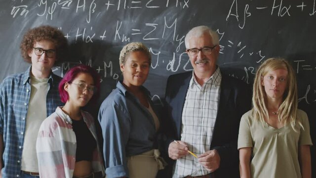 Waist up shot of senior professor and group of young diverse male and female students standing against chalkboard, smiling and posing together for camera in college classroom