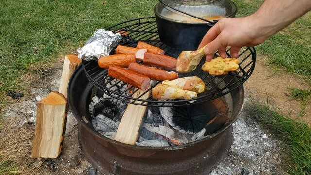Placing marinated chicken drumstick onto grill to be cooked over fire pit along side sausage and cast iron pot of soup.