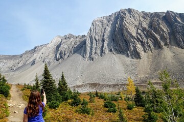 A young woman hiking and taking a selfie photo by a beautiful trail with a huge mountain in the background during a day in autumn, along the Ptarmigan Cirque trail in Kananaskis, Alberta, Canada