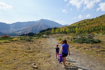 Fototapeta premium A mother and daugher hiking a beautiful trail above the treeline with a huge mountain in the background during a sunny day in autumn, along the Ptarmigan Cirque trail in Kananaskis, Alberta, Canada