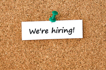 We're hiring. Text written on a piece of paper, cork board background.