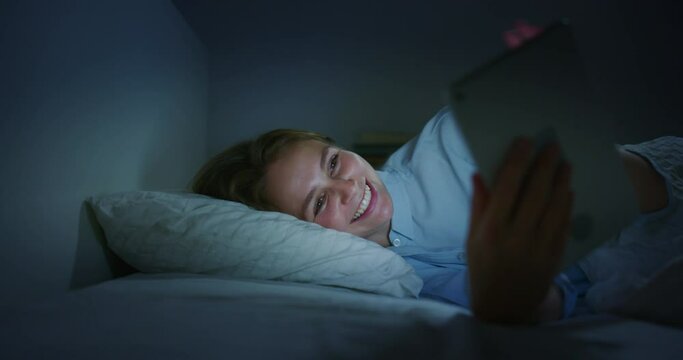 Authentic shot of an young blond woman is making a video call or selfie with a tablet before fall asleep in a cosy bed in a bedroom at night. Concept of technology, new generation,family, connection, 