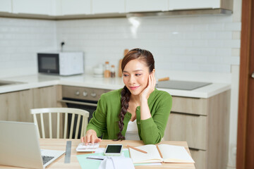 Attractive young smiling Woman paying bills online using laptop from home while sitting in the kitchen