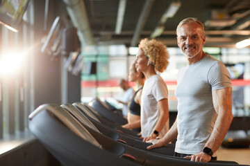 Portrait of mature man smiling at camera during his training on treadmill with other people...