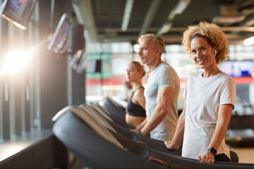 Portrait mature woman smiling at camera during her training on treadmill with other people in the background