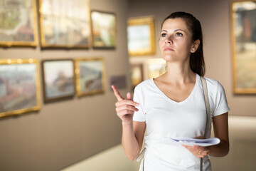 Focused adult girl holding brochure with exhibition program admiring paintings in museum