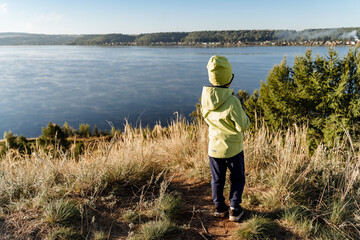 A little boy in a jacket walks along the lake in nature. The child explores the world around them....