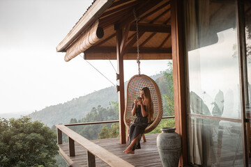 Tourist woman swing on wicker rattan hang chair in the jungle, nature mountains view, hold in hands...
