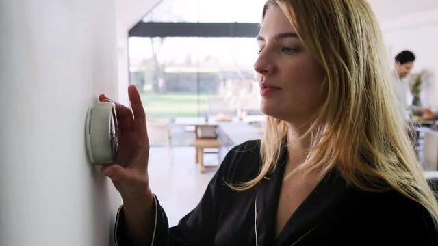Woman wearing pyjamas walks into shot and adjusts wall mounted digital central heating thermostat control at home - shot in slow motion