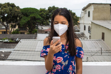Ni&ntilde;a latina con protecci&oacute;n mascarilla con celular desde su balc&oacute;n