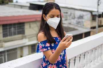 Ni&ntilde;a latina con protecci&oacute;n mascarilla con celular desde su balc&oacute;n