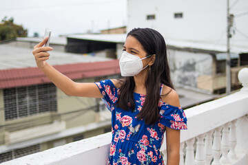 Ni&ntilde;a latina con protecci&oacute;n mascarilla con celular desde su balc&oacute;n