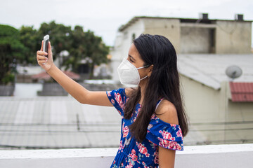 Ni&ntilde;a latina con protecci&oacute;n mascarilla con celular desde su balc&oacute;n