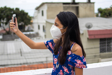 Ni&ntilde;a latina con protecci&oacute;n mascarilla con celular desde su balc&oacute;n