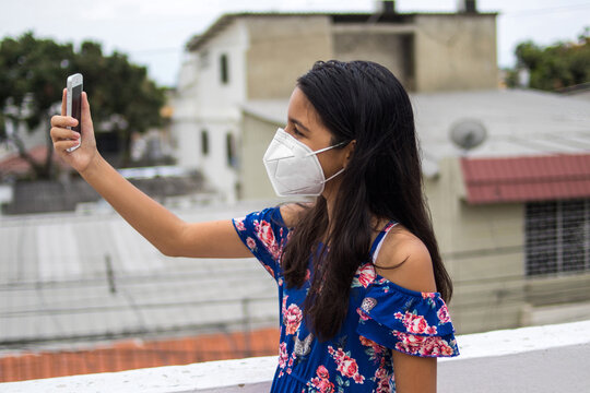 Niña latina con protección mascarilla con celular desde su balcón