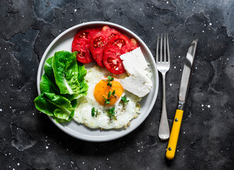 Breakfast, snack, tapas - fried egg, ripe red tomatoes, greek sheep cheese, green salad on a dark background, top view