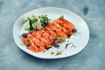 Sliced lightly salted salmon with arugula and parmesan. Red fish sashimi with capers and olives on a white plate on a gray background.. Fresh seafood