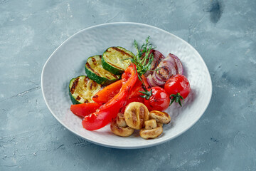 Grilled zucchini, bell peppers, onions, tomatoes and mushrooms in a white bowl. Vegetarian dish grilled vegetables on gray background