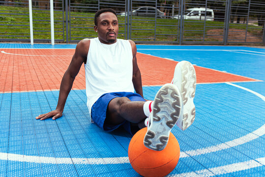 Young african american basketball player in white t-shirt and blue shorts sitting on basketball court put his feet on orange basketball, sports concept, healthy movement