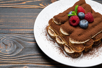 portion of Classic tiramisu dessert with raspberries and blueberries on ceramic plate on wooden background