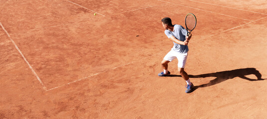 Tennis player playing on clay court. Young male tennis player hitting backhand kick. Professional athlete is concentrating in the game. Sports background. Banner. Copy space for text - obrazy, fototapety, plakaty