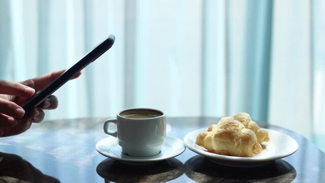 Closeup view of woman holding modern smartphone and taking pictures of her morning breakfast coffee and croissants to share photos on  social media resources
