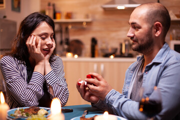Man celebrating relationship and asking girlfriend to marry him while having dinner. Man asking his girlfriend to marry in the kitchen during romantic dinner. Happy caucasian woman smiling being