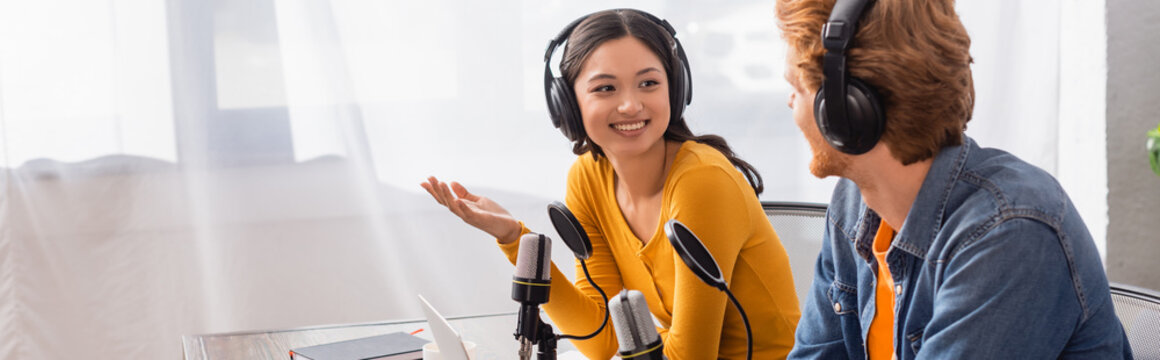 horizontal image of asian announcer in wireless headphones gesturing while talking to colleague in studio