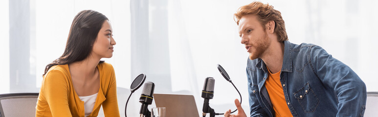 Obraz premium young redhead broadcaster talking to brunette asian woman during interview, panoramic shot