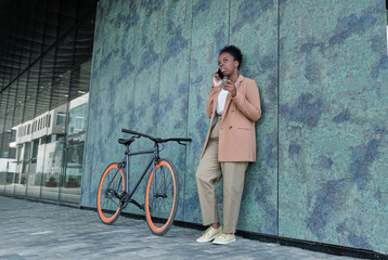 African young woman leaning on the wall of building talking on mobile phone during her ride on a bike