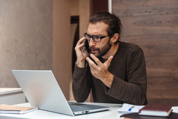 Portrait of serious man with beard talking on mobile phone and working at laptop at home