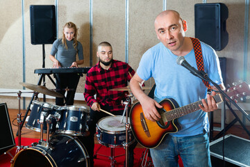 Rehearsal of music group. Cheerful band leader playing guitar and singing with his musicians in recording studio